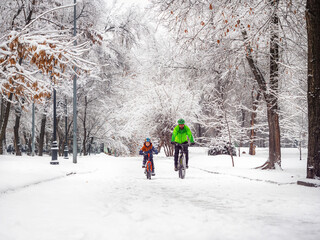 Father and son ride bicycles in a winter park. The trees around are beautifully covered with fluffy snow. Active family weekend in winter