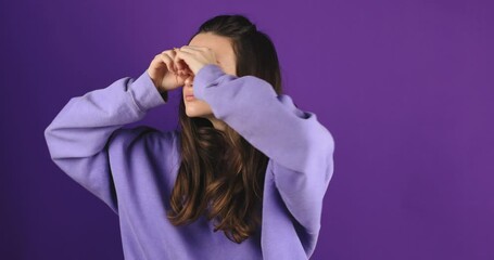 Amazed curious woman with brunette hair in violet hoody looking around through fingers imitating binoculars and point at something surprised. Indoor studio shot isolated on purple background.