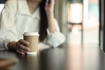 Businesswoman holding paper cup of hot drink in hands and talking on phone inside office building