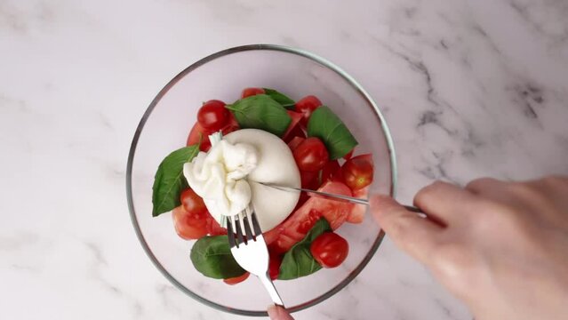 Burrata with tomatoes and basil salad, classic Italian dish, hands cutting a pouch of creamy stracciatella cheese