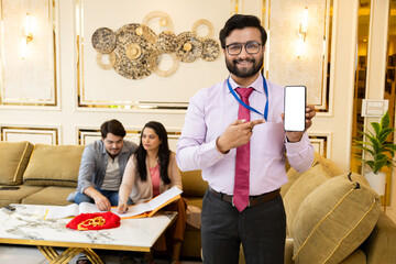 Happy looking indian Bank Agent showing mobile, smart phone. couple doing documentation process for gold loan in background with gold jewelery. Finance and Loan Concept. - obrazy, fototapety, plakaty