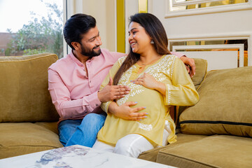 Happy indian couple expecting a baby while husband putting his hand on wife his pregnant wife belly both sitting on couch spending time together. healthcare concept