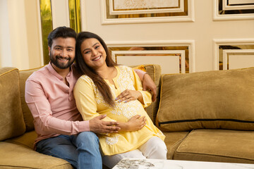 Happy indian couple expecting a baby while husband putting his hand on wife his pregnant wife belly both sitting on couch spending time together. healthcare concept