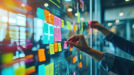  A professional's hands are arranging colorful sticky notes on a glass wall for project management and brainstorming in a modern office. © Rattanathip