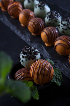 Handmade round shaped chocolates. A black plate made of stone. Belgian chocolate of the highest quality. Mint leaves are out of focus. Dessert for the holiday. Black background.Vertical photo.