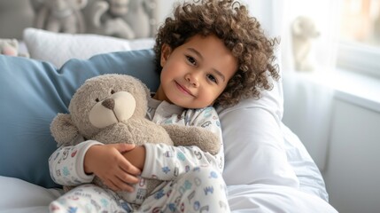 A young child with curly hair wearing pajamas holding a teddy bear and sitting on a bed with a blue pillow and a white blanket.