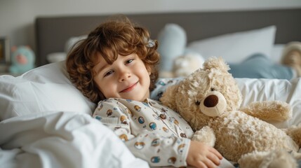 Young child with curly hair smiling and hugging a large teddy bear lying in a bed with white sheets and pillows surrounded by stuffed animals.