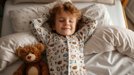 Fototapeta premium A young child with curly hair wearing patterned pajamas smiling and resting their head on a pillow with a teddy bear beside them on a bed.