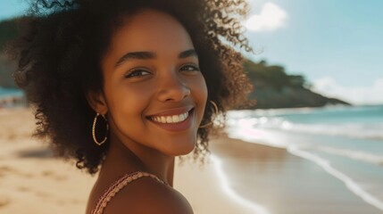 Smiling woman with curly hair wearing gold hoop earrings standing on a sandy beach with the ocean in the background.