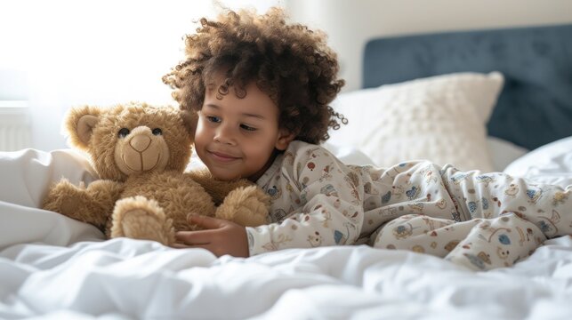Young child with curly hair wearing pajamas lying on bed with white sheets holding a brown teddy bear smiling and looking at the camera.