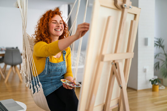 Satisfied young female artist sitting in her atelier and painting. Smiling woman holding color palette and paint brush, creating the art.