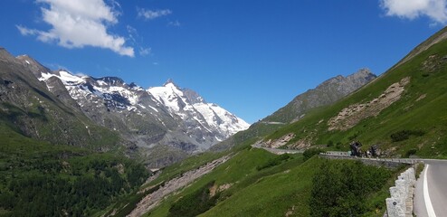 Scenic view of a road in green mountains with snow-covered peaks on a sunny day