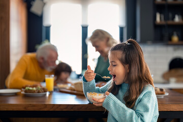 Selective focus of adorable small girl eating corn flakes cereals for breakfast while standing in...