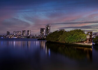 Naklejka premium Night-time scene of a river with a rusty ship with trees and a cityscape illuminated in the back