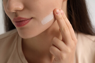 Young woman with dry skin applying cream onto her face on blurred background, closeup