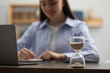 Hourglass with flowing sand on desk. Woman taking notes while using laptop indoors, selective focus