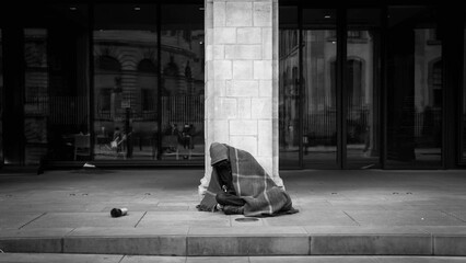 Homeless man sitting on a city street, covered in a blanket for warmth in grayscale