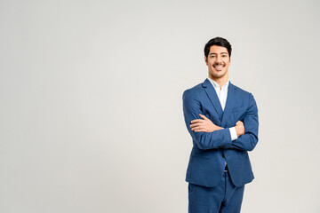 Cheerful young professional in smart blue suit with crossed arms showcases confidence and a readiness for business challenges, set against clean, light background that accentuates his sharp appearance
