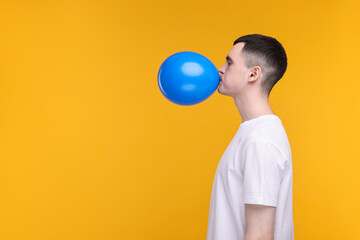 Young man inflating light blue balloon on yellow background. Space for text