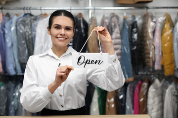 Dry-cleaning service. Happy worker holding Open sign at counter indoors, space for text