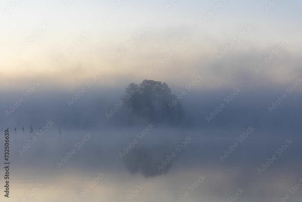 Poster Tranquil lake surrounded by a lush green landscape with fog rolling in from the horizon.