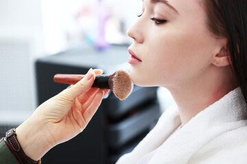 Close-up of the makeup artist's hand with a large powder brush. A beautiful young woman applies...