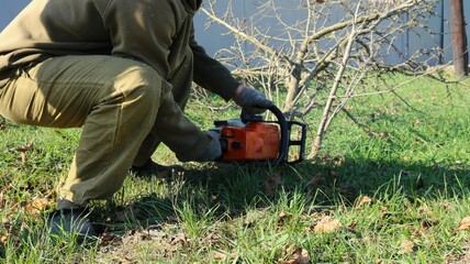 A man in a working dark green clothing with an orange chainsaw cuts a branchy bush on a solar lawn, cleaning the adjacent territory and getting rid of shrub vegetation using a gasoline saw