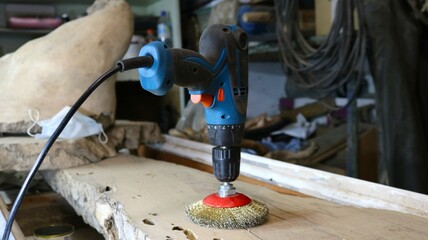 The wood processor tool, standing on a wooden workbench in the workshop, is a blue drill with a red round nozzle with golden metal bristles on a thick piece of a whole tree in the room
