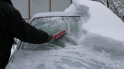 a person in black clothes cleans the window of a gray car from heavy snow with a brush after a snowstorm or blizzard with snowfall, eliminating the consequences of bad weather in the winter season