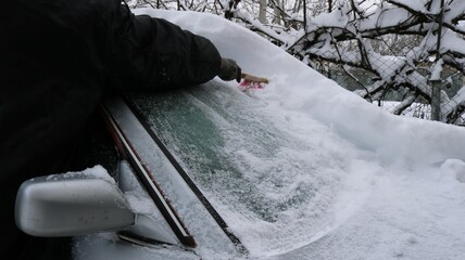 Person in warm black clothing clears a layer of snow from the windshield of a gray machine after winter bad weather, cleaning the car from snowy sediment manually after a plentiful snowfall