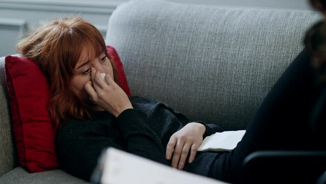 Unhappy young lady crying and talking to her personal psychologist. Psychotherapist appointment. Distressed woman lying on pillow and using napkin when crying. Caregiver listening grief story from