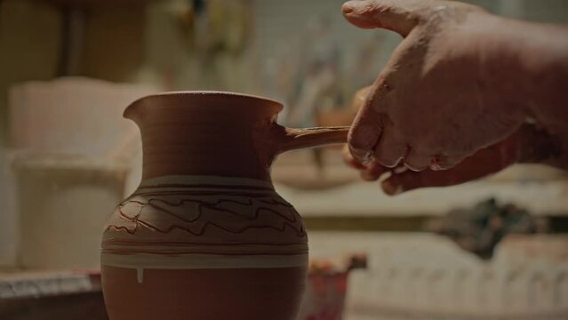 Close up potter's hands attaches hadle to clay jug and strengthens its form. Male professional potter working with extra details his handmade crockery. Old craftsman making ceramic pot in workshop