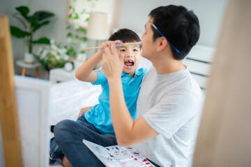 Young Asian dad playing with his son while teaching him painting and drawing on canvas at home with fun and smile of happiness. Father and kid relationship concept.