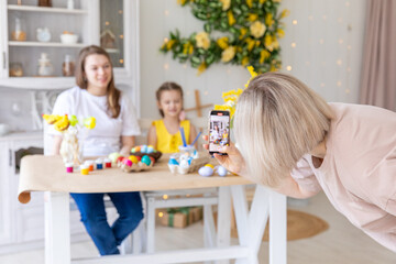 Obraz na płótnie Canvas Mom takes pictures of two daughters sitting at kitchen at home during Easter holidays. Easter preparation coloring eggs together taking selfie photo on smartphone smiling cheerful