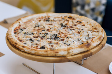 A large round fresh hot delicious pizza stands on a wooden stand in a pizzeria on the table. Food...