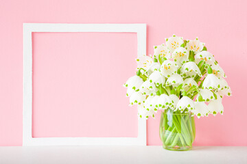 Bouquet of fresh beautiful snowdrops in vase on table at light pink wall background. Pastel color. First messengers of spring. Empty place for inspirational text in white frame. Front view. Closeup.