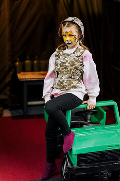 Beautiful girl, child stands in military camouflage, khaki uniform, helmet with face painted with strong tiger makeup near children's car. Photography, Ukrainian pariot, concept.