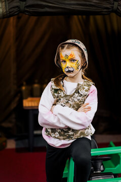 Beautiful girl, child stands in military camouflage, khaki uniform, helmet with face painted with strong tiger makeup near children's car. Photography, Ukrainian pariot, concept.