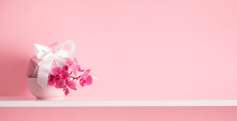 Gift box with and pink orchid on white shelf and on background of  pink wall. Greeting card for...