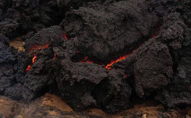 The quickly cooled lava crust creates an abstract. orange bright molten lava flowing on gray lava field and glossy rocky ground near  volcano with fog in background.