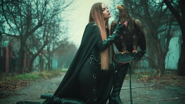 art portrait fantasy woman warrior holding white-tailed eagle wild bird on hand. Princess girl long blond hair. Lady queen assassin hunter black dress armor walking along street dark trees fog. 4k