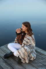 Fototapeta premium the girl is sitting on a wooden pier near the lake wrapped in a blanket and drinking coffee. aesthetic photo. autumn vibe