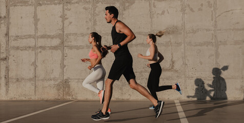 Three confident young people in sportswear running in front of the concrete wall outdoors together