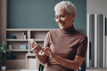 Senior businesswoman using smart phone and smiling while leaning at the office desk