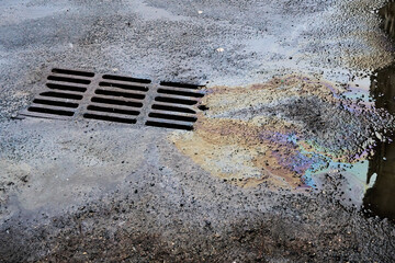 An oil or gasoline slick against the backdrop of an asphalt road flows into a storm drain....