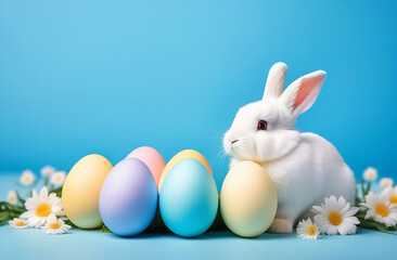 A small white fluffy rabbit sits near colorful Easter eggs and flowers on a blue background
