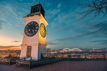 sun sets over Novi Sad, the iconic clock tower of Petrovaradin Fortress stands tall, offering a picturesque view of Serbian history and culture. - obrazy, fototapety, plakaty