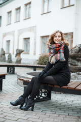 Happy beautiful European young girl with a sweet smile in a black fashion coat with a vintage checkered scarf, tights and shoes sits on a bench in the city