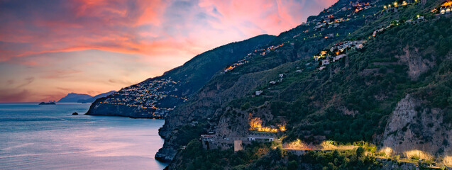 Amalfi Coast, Italy. View over Praiano on the Amalfi Coast at sunset. Street and house lights at dusk. In the distance the island of Capri on the horizon. Sea landscape. Banner header image.