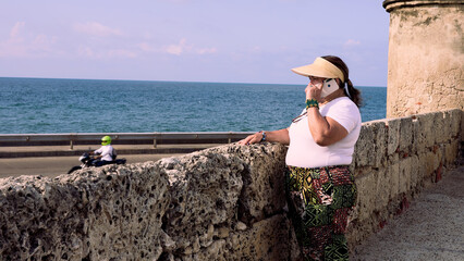 An elderly woman standing at the edge of the Cartagena city walls, dressed casually with a visor,...